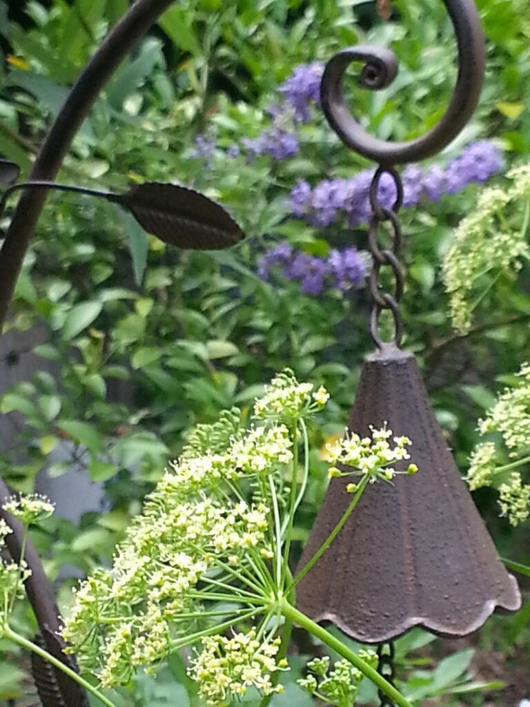 A rustic bell hanging among green foliage and purple flowers.